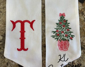 May include: Two white fabric items with red embroidery. One features a stylized letter "T", the other a Christmas tree in a red pot with a bow. The text "KL Print Shop" is at the bottom.