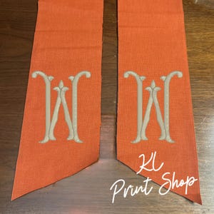 May include: Two orange fabric banners with embroidered gray capital "W" monograms. The banners have a pointed bottom edge. The text "KL Print Shop" is written in white script on the bottom right banner.