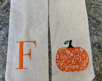 May include: Two beige kitchen towels. One towel has an embroidered orange letter "F". The other towel has an embroidered orange pumpkin with white decorative swirls and a green stem. The towels are made of a textured fabric.