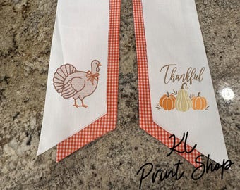 May include: Two white fabric table runners with orange and white gingham trim. One runner features an embroidered brown turkey with a bow. The other runner displays the word Thankful in brown script above three embroidered pumpkins in orange and tan.