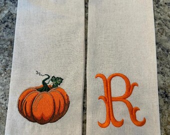 May include: Two off-white kitchen towels. One towel features an embroidered orange pumpkin with green leaves and a curly vine. The other towel has an embroidered orange letter "R".