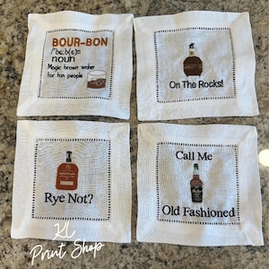 May include: Four white linen coasters with black stitched borders. Each coaster features a different bourbon-themed design with text and illustrations. The designs include "Bourbon" definition, "On The Rocks!", "Rye Not?", and "Call Me Old Fashioned".