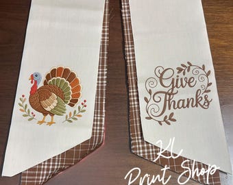 May include: Two off-white fabric table runners with brown and white checkered trim. One runner features an embroidered turkey with colorful feathers and foliage. The other runner displays the brown text Give Thanks surrounded by a decorative vine design.