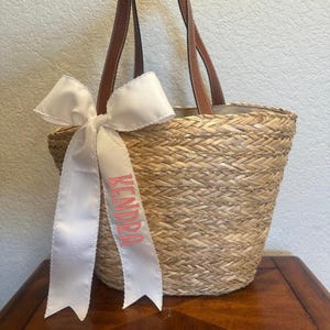 May include: A straw tote bag with brown leather handles and a white ribbon bow. The ribbon has the name "KENDRA" embroidered on it in pink.