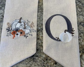 May include: Two beige fabric panels with embroidered designs. One panel features a white pumpkin with brown stems and orange and white flowers. The other panel features a gray letter O with a white pumpkin inside.