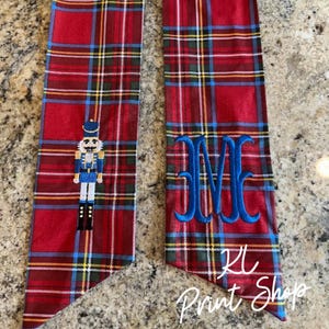 May include: Two red plaid fabric items. One features a nutcracker design, the other has blue embroidered initials. The text "KL Print Shop" is at the bottom of the second item. The plaid includes blue and yellow accents.