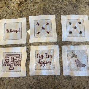 May include: Six white square embroidered coasters. Designs include the text "Whoop!", the letters "ATM", the text "Gig 'Em Aggies", footballs, dogs, and a dog with a maroon blanket. Each coaster has a dotted border.