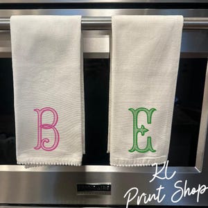 May include: Two white kitchen towels with embroidered letters, one pink "B" and one green "E". The towels hang from a metal bar. The towels have a textured pattern and a decorative edge. The text "KL Print Shop" is in the bottom right corner.
