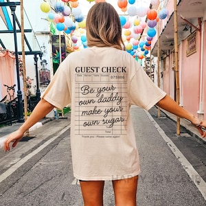 Op de afbeelding: Een beige t-shirt met een zwarte afbeelding van een gastenbon waarop staat "Be your own daddy make your own sugar".