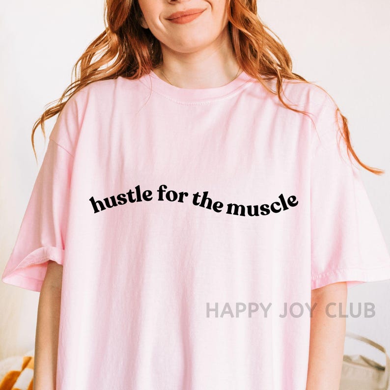 May include: Pink t-shirt with the text "hustle for the muscle" in black.