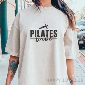 Op de afbeelding: Lichtbeige T-shirt met de zwarte tekst "PILATES babe". Het woord "PILATES" staat boven het woord "babe" en is onderstreept. Een silhouet van een persoon die pilates doet, staat boven het woord "PILATES".