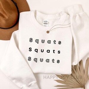 May include: White crewneck sweatshirt with the text "Squats" repeated three times in black outlined letters.