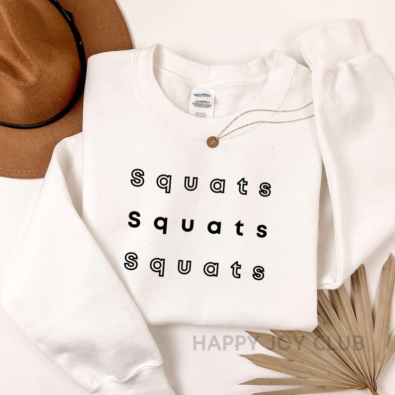 May include: White crewneck sweatshirt with the text "Squats" repeated three times in black outlined letters.
