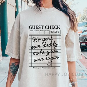 Op de afbeelding: Een beige t-shirt met een zwarte afbeelding van een gastenbon waarop staat "Be your own daddy make your own sugar". De afbeelding is op een witte achtergrond met zwarte lijnen.
