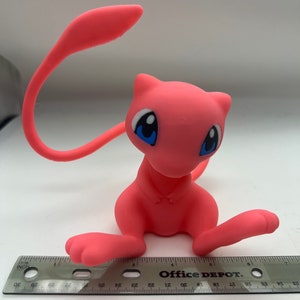 May include: Pink plastic figurine of a Pokemon character, Mew, with blue eyes. The figurine is approximately 4 inches tall and is sitting on a ruler.