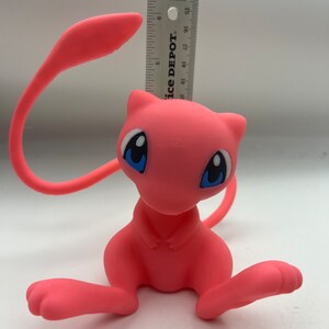 May include: A pink and white Pokemon figurine of a sitting  Mewtwo with blue eyes. The figurine is about 6 inches tall.