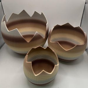 May include: A set of three round bowls with jagged, broken-egg-shell style rims. The bowls feature a horizontal gradient pattern in shades of brown, tan, cream, and soft blue. The surfaces have a fine, ribbed texture throughout the exterior.