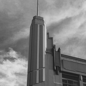 May include: Black and white photograph of a tall building with an Art Deco design. The building has a vertical tower with a flagpole at the top. The sky is overcast with dark clouds.