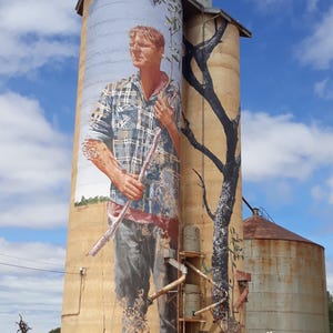 Peut inclure: Un grand silo à grain avec une peinture murale sur le côté. La peinture murale représente un homme en chemise à carreaux tenant une branche. Le silo est peint dans des tons de brun, beige et bleu. Le ciel est bleu avec des nuages blancs. Le texte "Faraway Photos" est visible en bas de l'image.