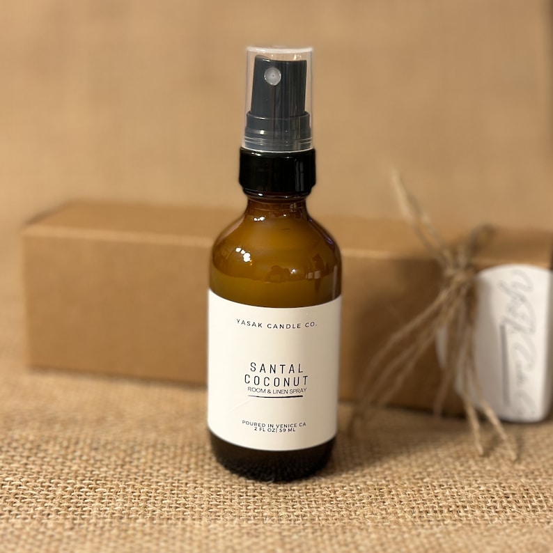 May include: A brown glass bottle with a black spray nozzle and a white label. The label reads "YASAK CANDLE CO. SANTAL COCONUT ROOM & LINEN SPRAY POURED IN VENICE CA 2 FL OZ / 59 ML".