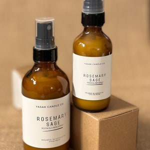 May include: Two amber glass bottles with black spray tops and white labels. The labels read "YASAK CANDLE CO. ROSEMARY SAGE ROOM & LINEN SPRAY" and include the size in ounces and milliliters. The bottles are sitting on a brown cardboard box.