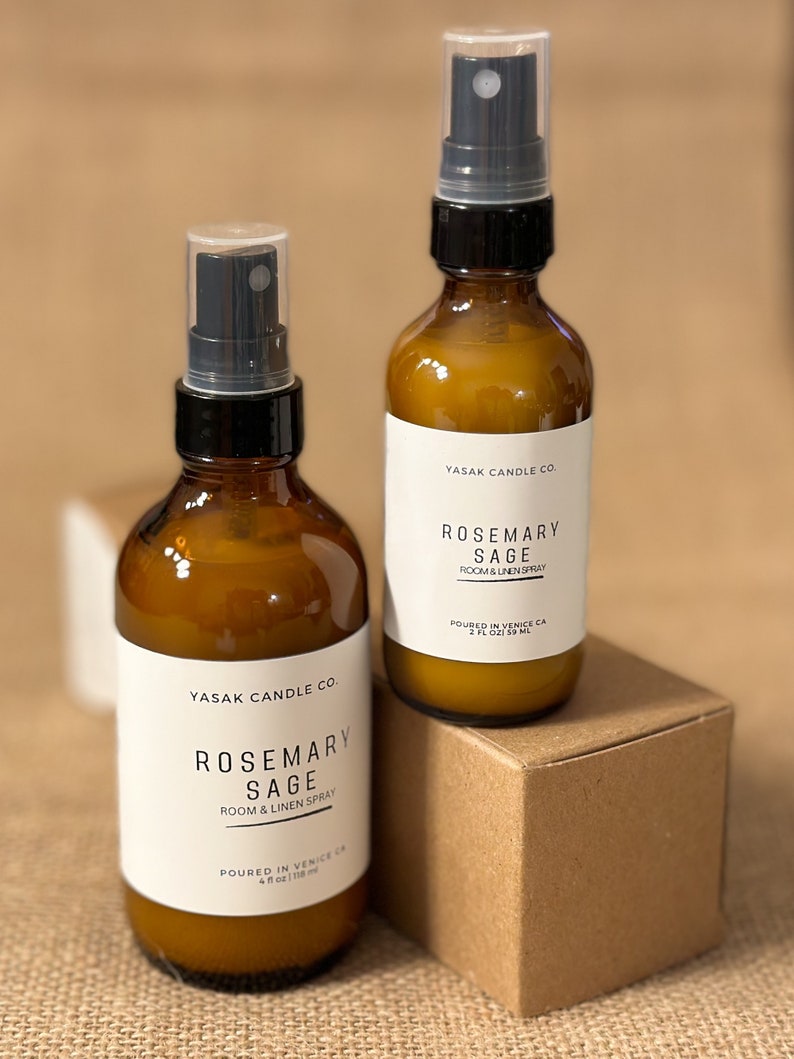 May include: Two amber glass bottles with black spray tops and white labels. The labels read "YASAK CANDLE CO. ROSEMARY SAGE ROOM & LINEN SPRAY" and include the size in ounces and milliliters. The bottles are sitting on a brown cardboard box.