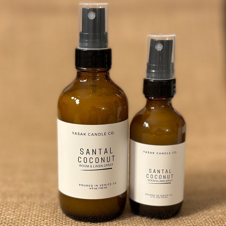 May include: Two amber glass bottles with black spray tops. Both bottles are labeled "YASAK CANDLE CO." and "SANTAL COCONUT ROOM & LINEN SPRAY". The larger bottle is labeled "POURED IN VENICE CA 4 fl oz | 118 ml". The smaller bottle is labeled "POURED IN VENICE CA 2 fl oz | 59 ml".