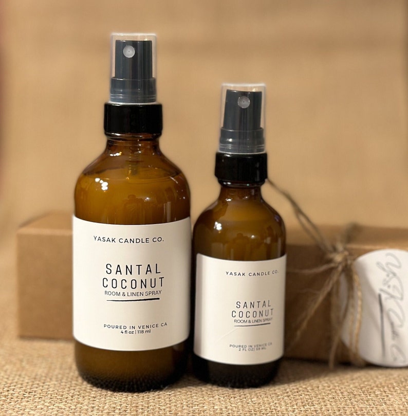May include: Two amber glass bottles with black spray tops and white labels. The labels read "YASAK CANDLE CO. SANTAL COCONUT ROOM & LINEN SPRAY". The larger bottle is labeled "4 fl oz 118 ml" and the smaller bottle is labeled "2 fl oz 59 ml".