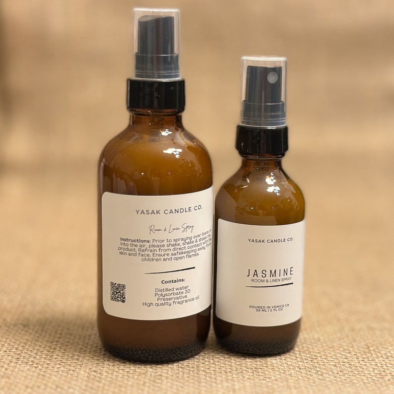 May include: Two amber glass bottles with black spray tops and white labels. The larger bottle is labeled "Yasak Candle Co. Room & Linen Spray" and the smaller bottle is labeled "Yasak Candle Co. Jasmine Room & Linen Spray".