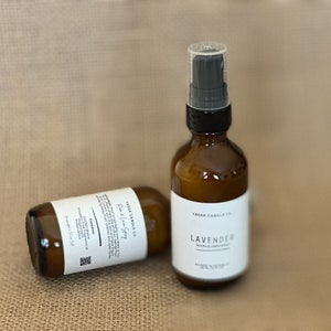 May include: Two amber glass bottles with black caps and white labels. The bottle on the left is labeled "YASAK CANDLE CO. Room & Linen Spray". The bottle on the right is labeled "YASAK CANDLE CO. LAVENDER Room & Linen Spray".