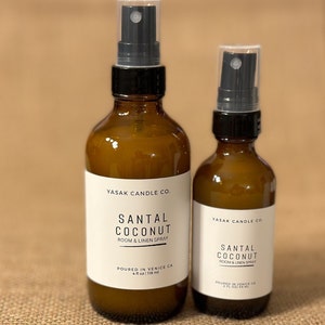 May include: Two amber glass bottles with black spray tops. The bottles are labeled "YASAK CANDLE CO." and "SANTAL COCONUT ROOM & LINEN SPRAY". The larger bottle is 4 fl oz / 118 ml and the smaller bottle is 2 fl oz / 59 ml.