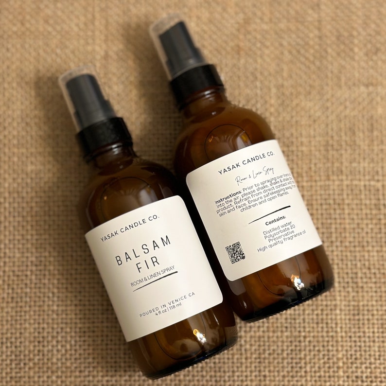 May include: Two amber glass bottles with black spray tops and white labels. The labels read "YASAK CANDLE CO." and "BALSAM FIR ROOM & LINEN SPRAY". The bottle on the right also includes instructions for use and a list of ingredients.
