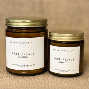 May include: Two amber glass candle jars with gold lids. The larger jar is labeled "YASAK CANDLE CO. ROSE PETALS SOY CANDLE HANDPOURED IN VENICE CA 7 oz | 198 g". The smaller jar is labeled "YASAK CANDLE CO. ROSE PETALS SOY CANDLE HANDPOURED IN VENICE CA 3.60oz | 102g".
