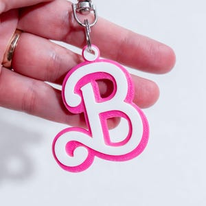 May include: A pink and white letter "B" keychain with a silver clasp. The letter is a layered design, with a white cursive "B" set against a pink background. The keychain is held up by a silver clasp.