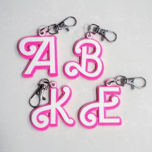 May include: Four keyrings featuring the letters A, B, K, and E. Each letter is white with a pink border and attached to a silver clasp. The keyrings are displayed on a white surface.