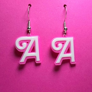 May include: Two earrings, each shaped like the letter "A" in a white, stylized font with a pink outline. They hang from silver-coloured fish hook ear wires, set against a bright pink backdrop.