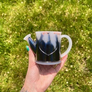 May include: A clear watering can-shaped mug with a heart design. The mug is filled with a dark liquid and has a white handle and spout. The mug is held in a hand against a blurred green background.