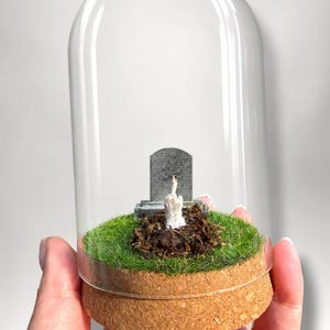 May include: A clear glass cloche dome sits on a circular cork base. Inside, a miniature grey tombstone stands on a patch of green artificial grass and dark brown soil. A small, white, hand-shaped figurine emerges from the soil, gesturing with a middle finger.