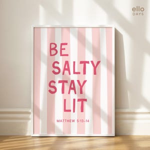 May include: A framed print with a pink and white striped background. Bold pink text reads "BE SALTY STAY LIT" with "MATTHEW 5:13-14" below. The artwork is in a white frame, suitable for home decor.