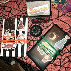 May include: Halloween-themed gift boxes and a black pumpkin on an orange spiderweb tablecloth. The boxes are decorated with stickers and wrapped with orange and green twine. One box reads "Monster Mash."