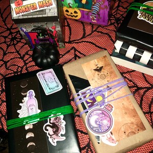 May include: Halloween-themed gift boxes and wrapped packages with stickers and twine. The boxes feature spooky illustrations and the text "Monster Mash." The wrapped gifts are adorned with stickers of moons, potions, and celestial designs.