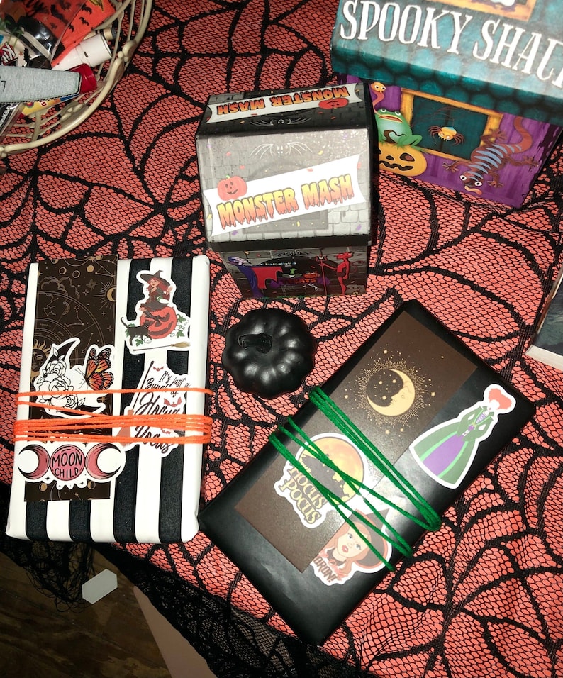 May include: Halloween-themed gift boxes and a black pumpkin on an orange spiderweb tablecloth. The boxes are decorated with stickers and wrapped with orange and green twine. One box reads "Monster Mash."