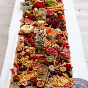 Peut inclure: Un long plateau rectangulaire de charcuterie rempli d'une variété d'aliments, notamment des fruits, du fromage, des craquelins, des noix et du salami. Le plateau est décoré de fleurs fraîches et est posé sur une table blanche.
