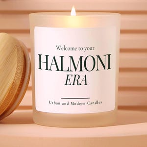 May include: A frosted glass candle with a lit wick and a wooden lid. The white label reads Welcome to your HALMONI ERA Urban and Modern Candles. Text on the right states No harmful chemicals like phthalates, parabens, or sulfates.