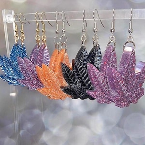Puede incluir: Seis pares de pendientes colgantes con un diseño de hoja de marihuana. Las hojas están hechas de resina y vienen en una variedad de colores, incluyendo azul, morado, naranja, negro y rosa.