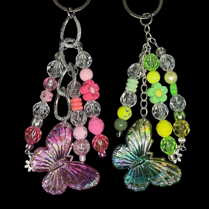Puede incluir: Dos llaveros de mariposa con cuentas. Uno presenta una mariposa rosa y morada con cuentas rosas y transparentes, y detalles de flores rosas. El otro tiene una mariposa verde y azul con cuentas verdes, amarillas y transparentes, y detalles de flores verdes.