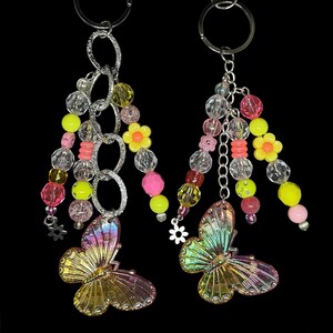 Puede incluir: Dos llaveros con eslabones de cadena plateados y detalles de cuentas de colores. Cada llavero presenta un colgante de mariposa con alas iridiscentes en tonos rosa, amarillo y verde. Las cuentas son rosas, amarillas, transparentes y naranjas.