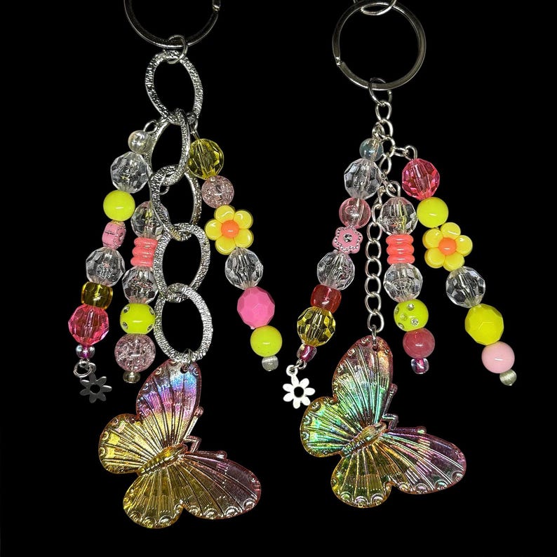 Puede incluir: Dos llaveros con eslabones de cadena plateados y detalles de cuentas de colores. Cada llavero presenta un colgante de mariposa con alas iridiscentes en tonos rosa, amarillo y verde. Las cuentas son rosas, amarillas, transparentes y naranjas.