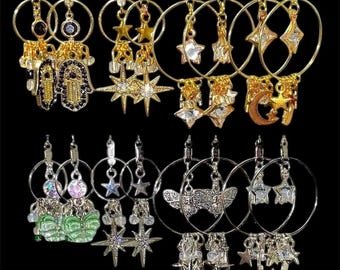 May include: A collection of eight pairs of hoop earrings arranged in two rows. The top row features gold-tone hoops with hamsa, star, and moon charms. The bottom row displays silver-tone hoops with green butterfly, star, moth, and moon charms.