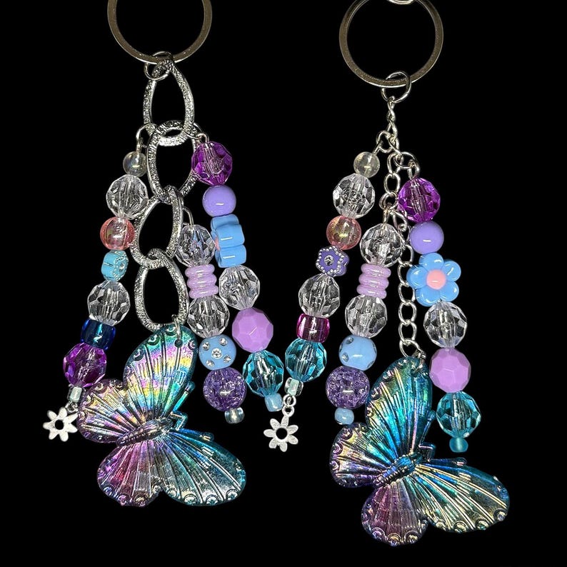 Puede incluir: Dos llaveros con colgantes de mariposa iridiscentes. Cada llavero presenta un anillo plateado, una cadena y hebras de cuentas en tonos morado, azul, rosa y transparente. Peque&ntilde;os dijes de flores acent&uacute;an el dise&ntilde;o sobre un fondo negro.